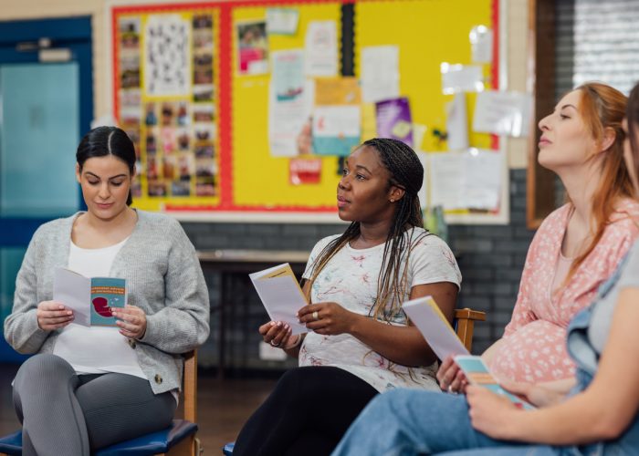 An over the shoulder view of a group of young pregnant women who are listening to a pregnancy fitness instructor and antenatal expert. She is showing them a leaflet and encouraging them to take part in a fitness class in a community centre in the North East of England.