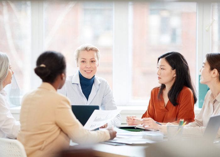 Portrait of diverse group of businesswomen discussing project while sitting at table during meeting in office, focus on smiling female boss